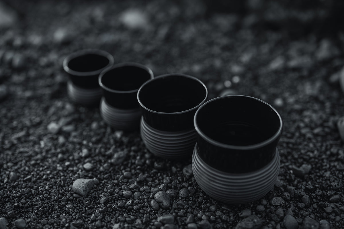 Four matte black porcelain cups arranged on dark volcanic gravel.