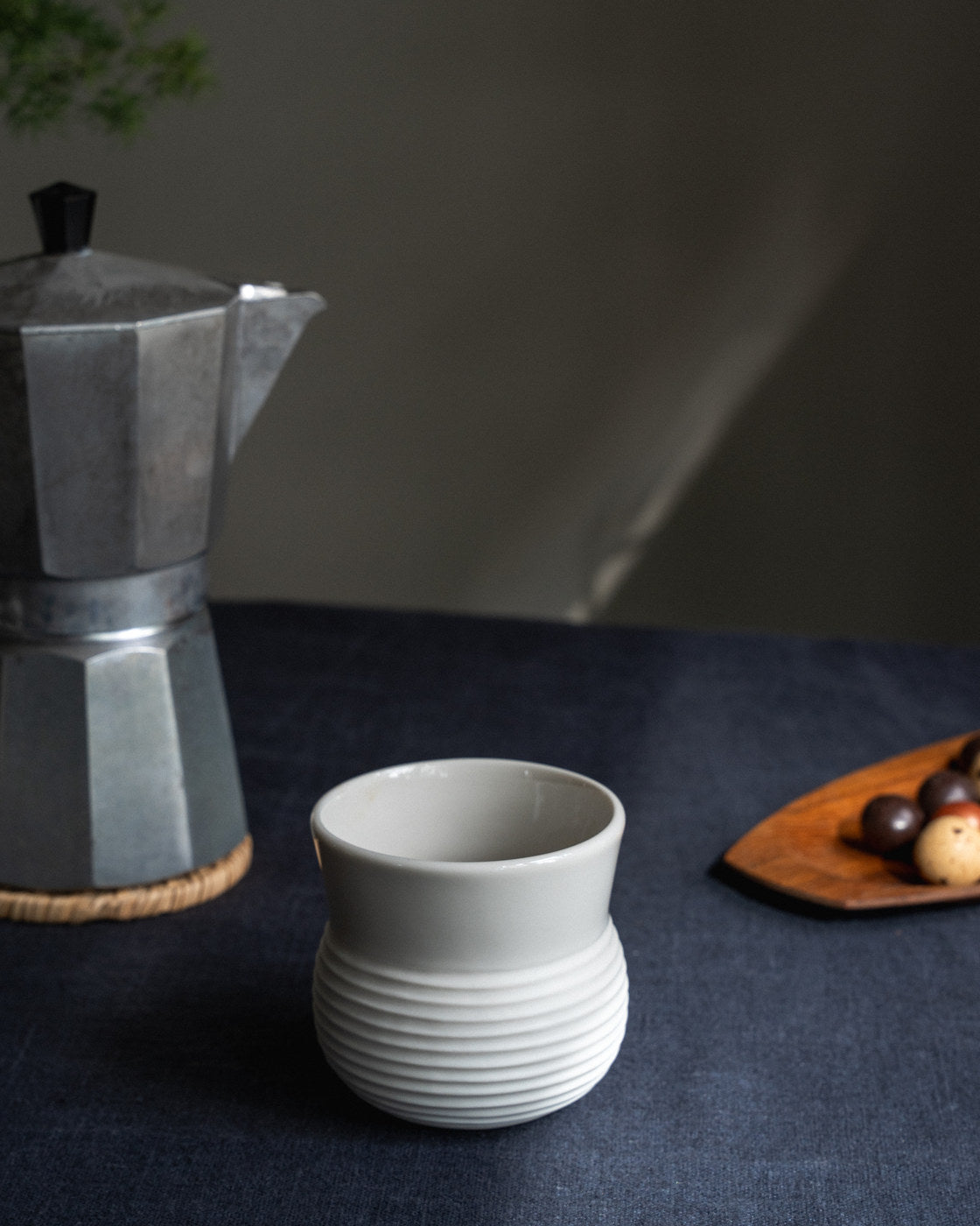 Minimal white porcelain cup beside a coffee maker on a blue linen tablecloth.